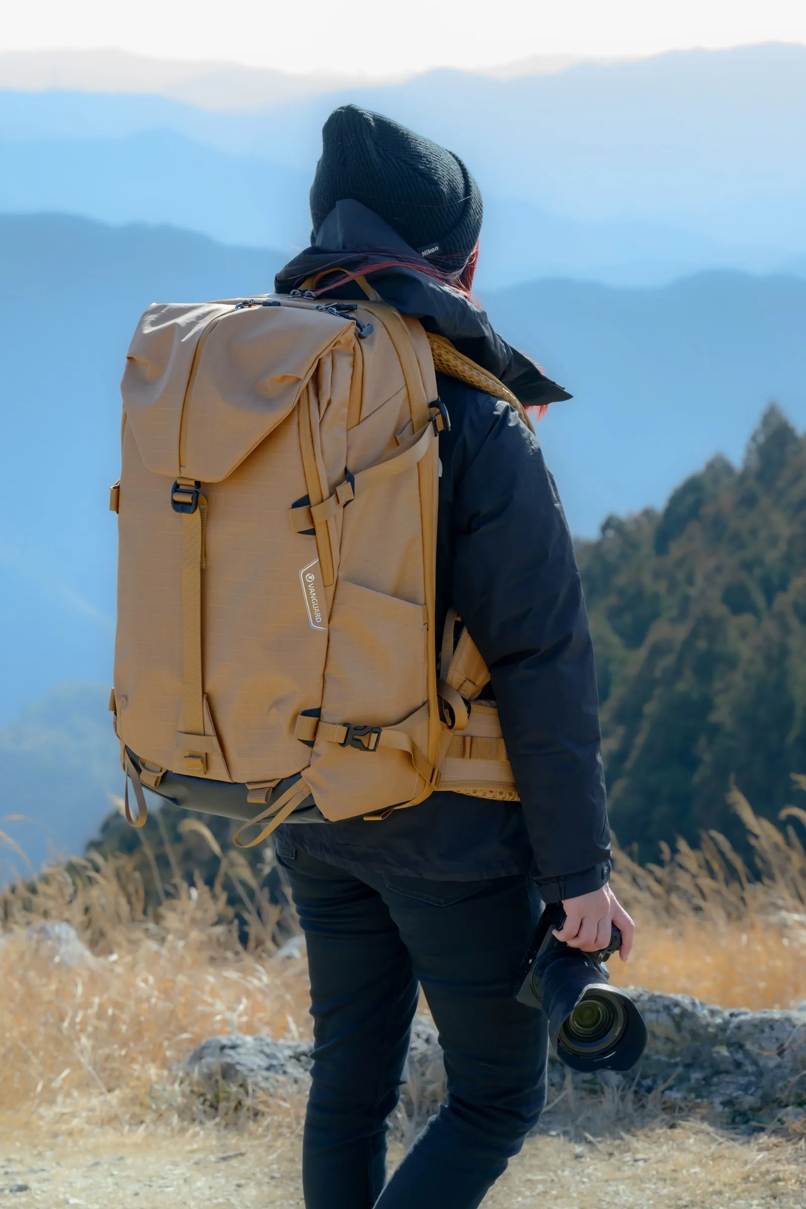 山の風景を前に黄色いカメラバッグを背負うモデル